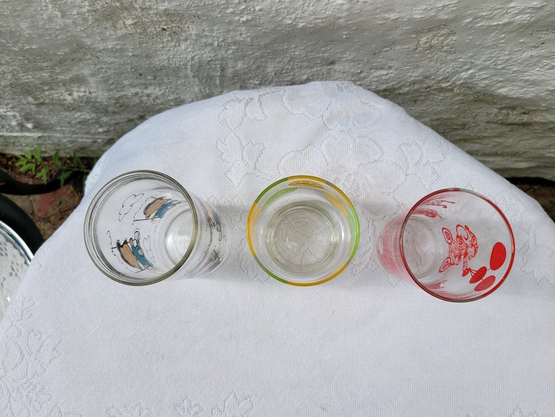 May include: Three vintage drinking glasses with colorful cartoon designs. The glasses are clear with a blue, yellow, and red rim. The designs are of a person in a boat, a person with a yellow umbrella, and a person with a red balloon.