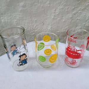 May include: Three glass drinking glasses with different designs. One glass has a yellow and green smiley face pattern, one has a red and white cartoon design, and one has a cartoon character design with blue, white, and black colors.