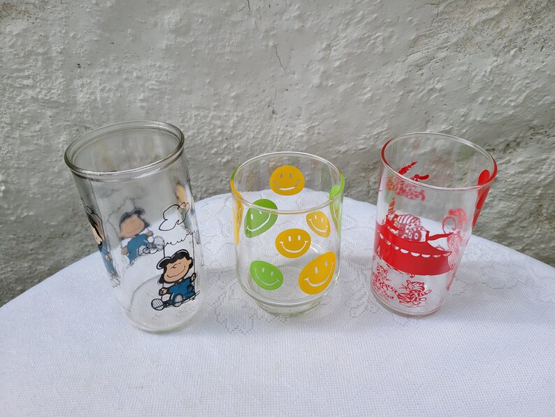 May include: Three glass drinking glasses with different designs. One glass has a yellow and green smiley face pattern, one has a red and white cartoon design, and one has a cartoon character design with blue, white, and black colors.