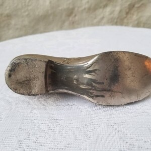 Pode incluir: Um objeto de prata em forma de sapato com uma superf&iacute;cie desgastada e texturizada. O sapato &eacute; um objeto decorativo, possivelmente um peso de papel ou uma pequena figura.