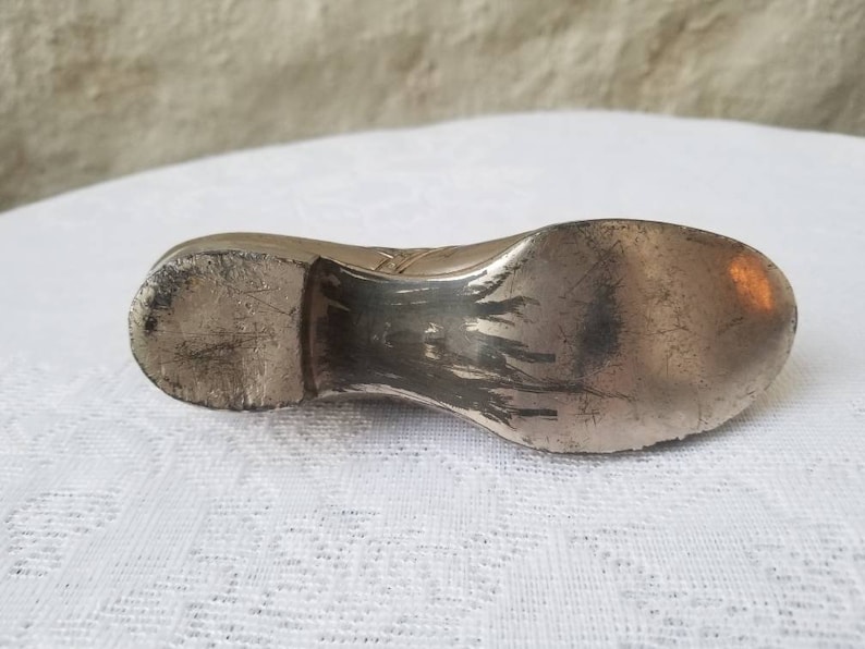 Pode incluir: Um objeto de prata em forma de sapato com uma superf&iacute;cie desgastada e texturizada. O sapato &eacute; um objeto decorativo, possivelmente um peso de papel ou uma pequena figura.