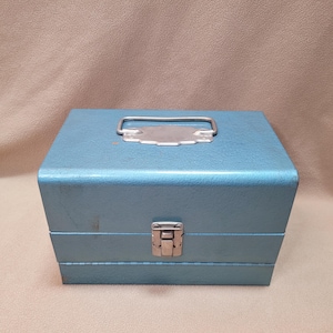 May include: A light blue metal toolbox with a silver handle and latch. The box is rectangular with a textured surface. The handle is attached to a silver plate on the top of the box.