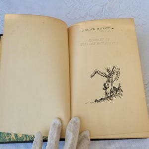 May include: An open book with a black and white illustration of a person standing next to a tree. The book's title page reads "Black Majesty" by William MacLeod Raine.