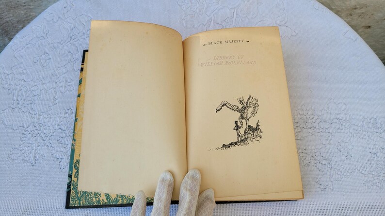 May include: An open book with a black and white illustration of a person standing next to a tree. The book's title page reads "Black Majesty" by William MacLeod Raine.