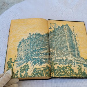 May include: An open book with yellowed pages and a black spine. The book features an illustration of a large, stone building with a green roof and a crowd of people surrounding it.
