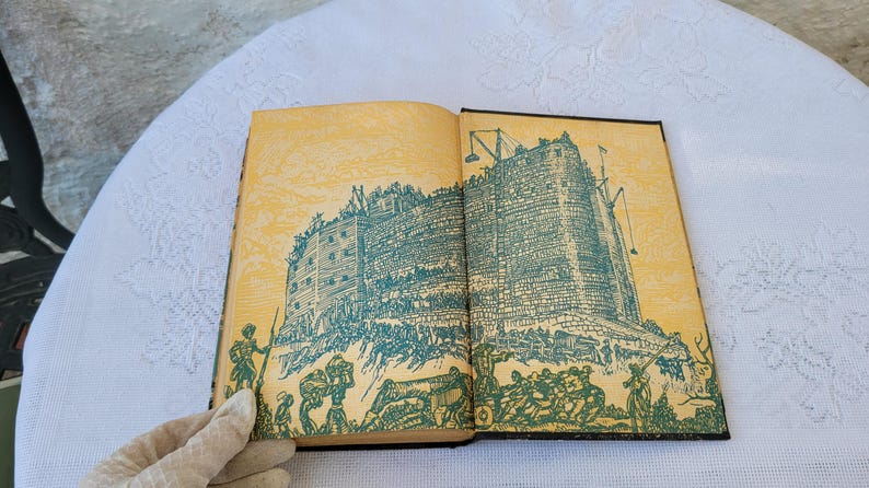 May include: An open book with yellowed pages and a black spine. The book features an illustration of a large, stone building with a green roof and a crowd of people surrounding it.