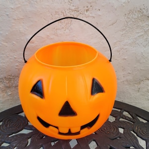 May include: Orange plastic pumpkin shaped trick-or-treat bucket with a black handle and a smiling jack-o'-lantern face.