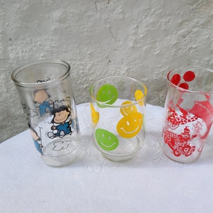 May include: Three clear glass drinking glasses with different designs. The first glass has a cartoon of Snoopy and Woodstock. The second glass has a yellow smiley face design. The third glass has a red design of a clown and balloons.