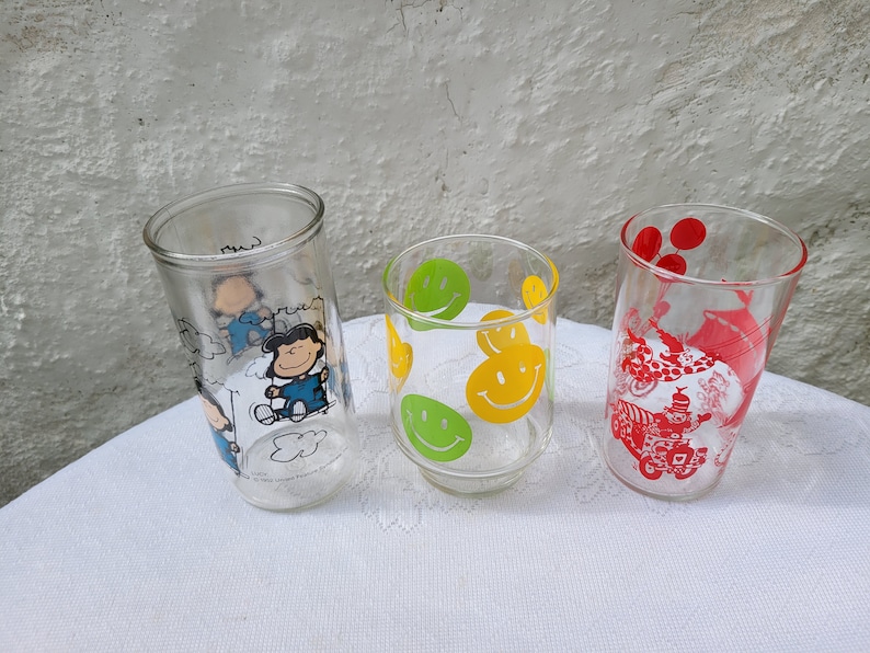 May include: Three clear glass drinking glasses with different designs. The first glass has a cartoon of Snoopy and Woodstock. The second glass has a yellow smiley face design. The third glass has a red design of a clown and balloons.