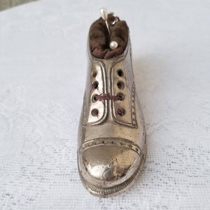 Pode incluir: Um almofadinha para alfinetes de metal prateado em forma de sapato com um interior de tecido marrom. O almofadinha tem um design de cadar&ccedil;o e est&aacute; cheio de alfinetes.