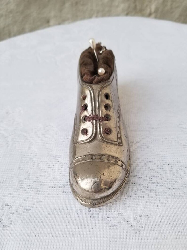 Pode incluir: Um almofadinha para alfinetes de metal prateado em forma de sapato com um interior de tecido marrom. O almofadinha tem um design de cadar&ccedil;o e est&aacute; cheio de alfinetes.