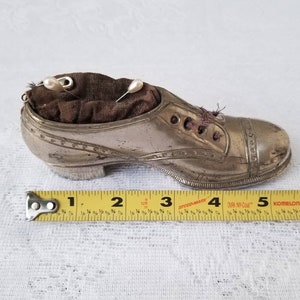 Pode incluir: Um almofadinha de alfinetes de prata em forma de sapato com um interior de tecido castanho e dois alfinetes com cabe&ccedil;a de p&eacute;rola branca. O sapato tem 12,7 cm de comprimento.