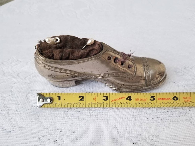 Pode incluir: Um almofadinha de alfinetes de prata em forma de sapato com um interior de tecido castanho e dois alfinetes com cabe&ccedil;a de p&eacute;rola branca. O sapato tem 12,7 cm de comprimento.