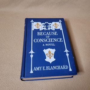 May include: A hardcover book titled "BECAUSE OF CONSCIENCE A NOVEL" by Amy E. Blanchard. The book has a blue cover with white decorative elements and text. The cover features a shield with a fleur-de-lis.