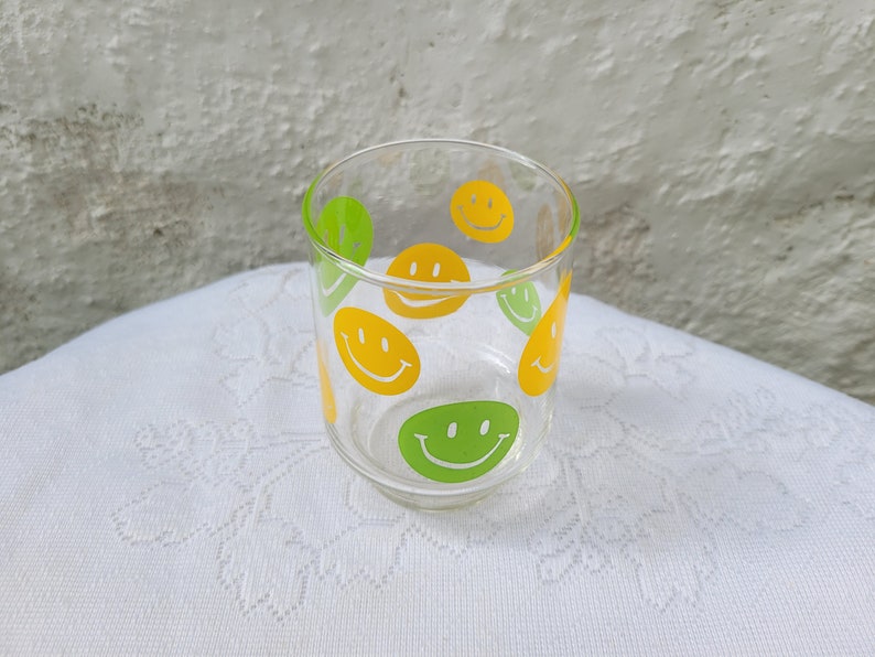 May include: A clear glass with yellow and green smiley faces printed on the side.