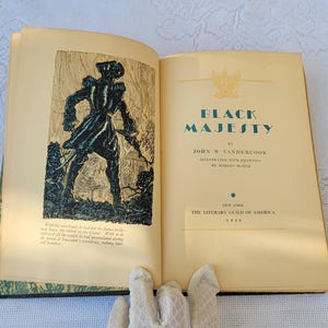 May include: An open book titled "Black Majesty" by John W. Vandercook. The book cover features a black and white illustration of a man in a military uniform standing in a forest.