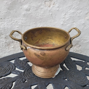 May include: A vintage copper bowl with two handles, resting on a decorative black metal surface. The bowl has a pedestal base and a golden interior, showing signs of age and use. The exterior has a reddish-brown patina.