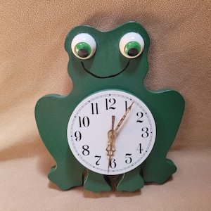 May include: A green frog-shaped wall clock with a white circular face featuring black Arabic numerals. The clock has two large, protruding white eyes with green pupils and a thin black line for a mouth. It includes ornate gold-colored hands.