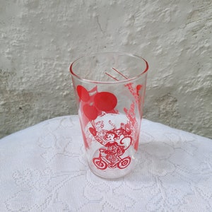 May include: A clear glass drinking glass with a red design of a monkey riding a tricycle with balloons.