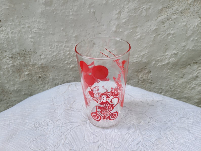 May include: A clear glass drinking glass with a red design of a monkey riding a tricycle with balloons.