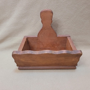 May include: A medium-brown wooden caddy featuring a rectangular base with a scalloped front edge and a tall, central vertical handle with a rounded top. The open-top container has straight sides and a smooth, stained wood finish throughout.