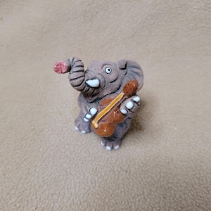 May include: A small, gray, textured elephant figurine holding a brown violin with a yellow neck. The elephant has white tusks, white toenails, and a pinkish tip on its trunk. The figure is depicted in a standing position with its trunk raised.