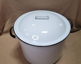 Puede incluir: Una olla de esmalte blanco con tapa a juego. La olla tiene asas negras y un borde negro. La tapa tiene un asa pequeña. La olla es adecuada para cocinar y tiene un aspecto clásico y vintage.