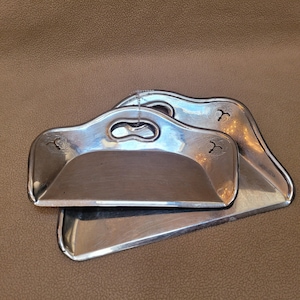 May include: Two silver-toned metal crumb scoops with integrated handles. Each scoop features a smooth, curved basin and decorative etched floral patterns on the sides. The scoops are stacked, showing a polished, reflective surface finish.