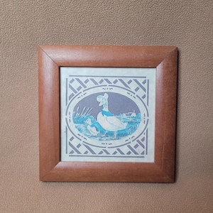 May include: A framed print featuring a duck and duckling in a rural scene. The artwork is in shades of blue and white, set within an oval frame, and surrounded by a decorative border. The wooden frame adds a rustic touch.