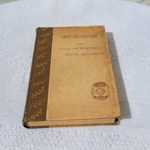 May include: A vintage hardcover book titled "The Vicar of Wakefield" by Oliver Goldsmith. The book has a brown cover with gold leaf accents and a decorative crest on the front.