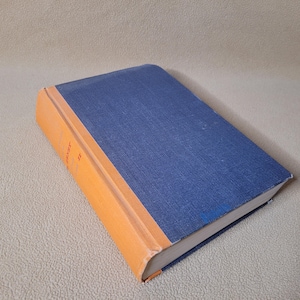 May include: A hardcover book with a blue cloth cover and an orange spine. The book's pages are white, and the spine has text in a dark color. The book is slightly angled, showing its cover and spine.