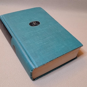 May include: A hardcover book with a teal cover and a black spine. The cover features a small, black circular emblem. The pages are aged, with a light tan color. The book is closed and resting on a neutral surface.