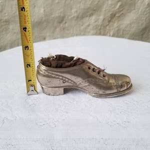 Pode incluir: Um almofadinha para alfinetes em forma de sapato prateado com um interior de veludo marrom escuro. O almofadinha para alfinetes mede aproximadamente 12,7 cm de comprimento.