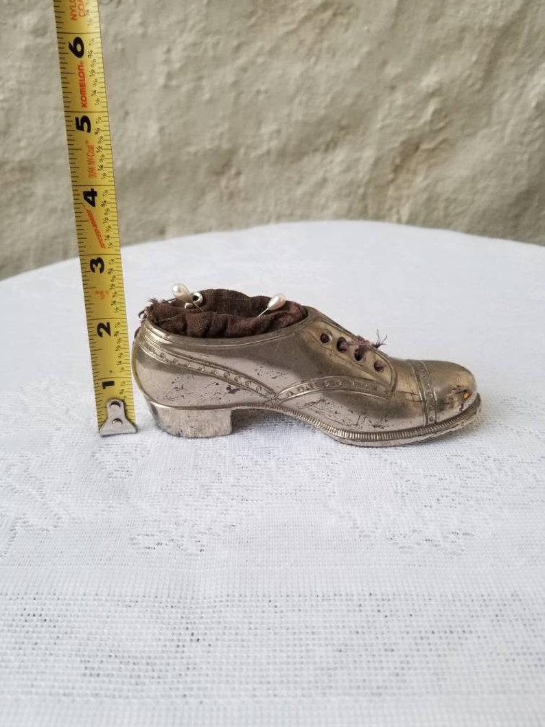Pode incluir: Um almofadinha para alfinetes em forma de sapato prateado com um interior de veludo marrom escuro. O almofadinha para alfinetes mede aproximadamente 12,7 cm de comprimento.