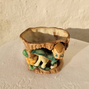 Puede incluir: Un macetero de cerámica con forma de tronco de árbol con un pequeño gnomo asomándose desde detrás del tronco. El gnomo lleva una camisa verde y pantalones marrones. El macetero está decorado con hojas verdes y setas amarillas.