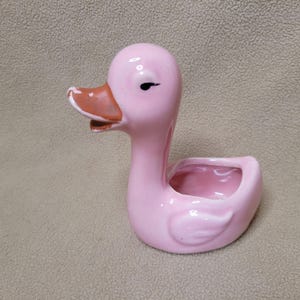 May include: A pink ceramic duck-shaped planter or trinket dish. The duck has a smooth, glossy finish, a brown beak with visible paint wear along the edges, and a simple black painted eye. The hollow body features an open top for storage.