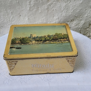 May include: A vintage yellow tin box with a picture of a river and buildings on the lid. The text "Milady Confectionery of Quality" is printed on the front of the box.