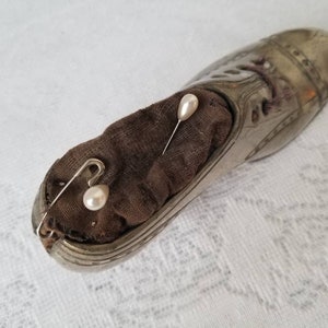 Pode incluir: Um almofadinha de alfinetes de prata em forma de sapato com um interior de tecido castanho. Dois alfinetes com cabe&ccedil;a de p&eacute;rola branca est&atilde;o presos no tecido.