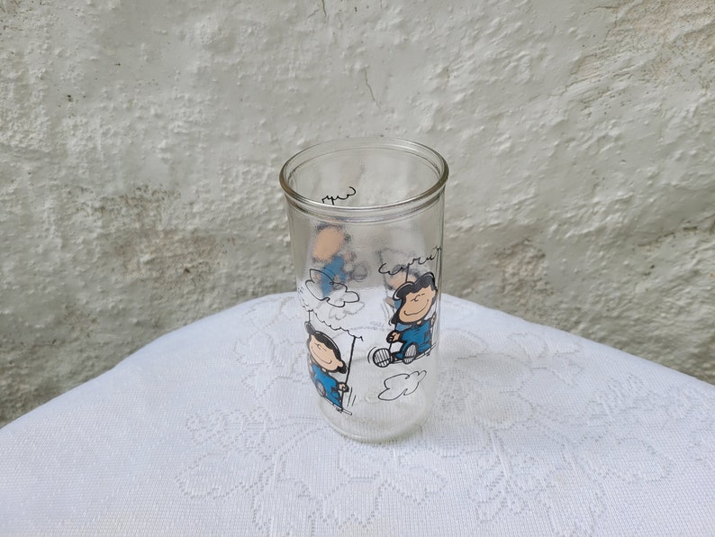 May include: A clear glass with a Peanuts cartoon design featuring Snoopy and Woodstock. The glass has a white background with blue and black cartoon characters.