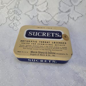 May include: A rectangular vintage Sucrets antiseptic throat lozenge tin with a cream-colored lid and dark blue base. The lid features the text Sucrets, Twenty-Four, and details about germicidal activity, Merck Sharp & Dohme, and Made in U.S.A.