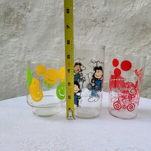 May include: Three vintage glass drinking glasses with different designs. The first glass has a yellow and green smiley face design. The second glass has a Peanuts cartoon design with Charlie Brown and Snoopy. The third glass has a red and white design with a cartoon character riding a motorcycle.