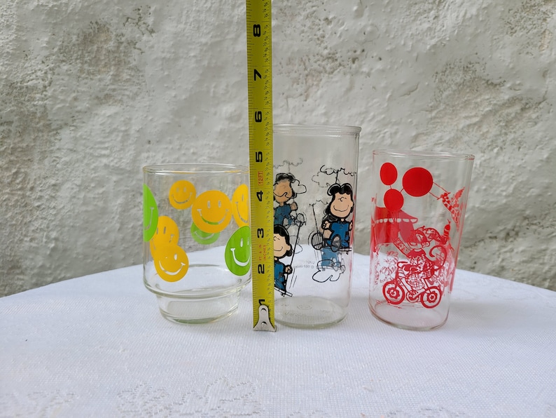 May include: Three vintage glass drinking glasses with different designs. The first glass has a yellow and green smiley face design. The second glass has a Peanuts cartoon design with Charlie Brown and Snoopy. The third glass has a red and white design with a cartoon character riding a motorcycle.