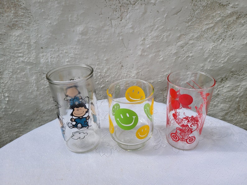 May include: Three glass drinking glasses with different designs. The first glass has a cartoon image of a character from the Peanuts comic strip. The second glass has a yellow smiley face design. The third glass has a red design of a person riding a bicycle with balloons.