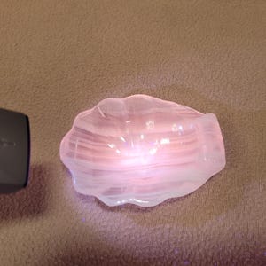May include: A translucent, pale pink, shell-shaped dish with a smooth, polished surface. The object features subtle, horizontal white banding throughout its structure and emits a soft, glowing light from within its center, highlighting its form.