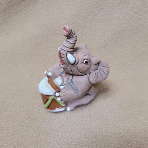 May include: A small, textured brown ceramic figurine of an elephant with its trunk raised upward. The elephant is depicted playing a drum with a white top, brown sides, and green accents. It features white tusks and detailed etched lines.
