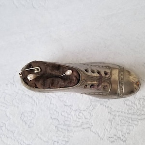 Pode incluir: Um almofadinha para alfinetes de metal prateado em forma de sapato com um interior de tecido castanho. O almofadinha para alfinetes tem um design de atacadores e dois alfinetes em forma de p&eacute;rola branca.