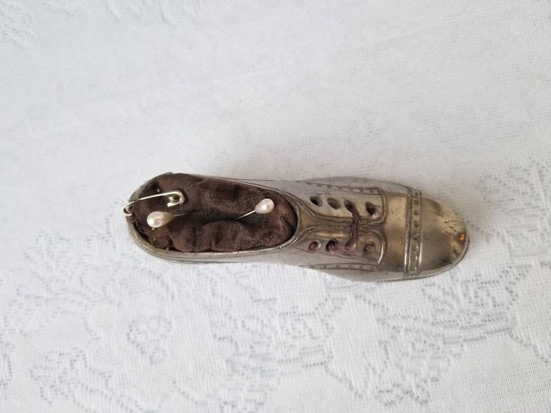 Pode incluir: Um almofadinha para alfinetes de metal prateado em forma de sapato com um interior de tecido castanho. O almofadinha para alfinetes tem um design de atacadores e dois alfinetes em forma de p&eacute;rola branca.