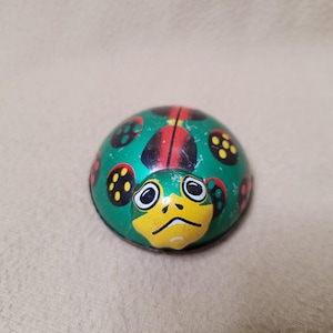 May include: A dome-shaped metal toy featuring a green base with a yellow face and black eyes. The shell is decorated with red and black spots, some containing yellow dots. The surface shows signs of wear with minor paint chips and scratches.