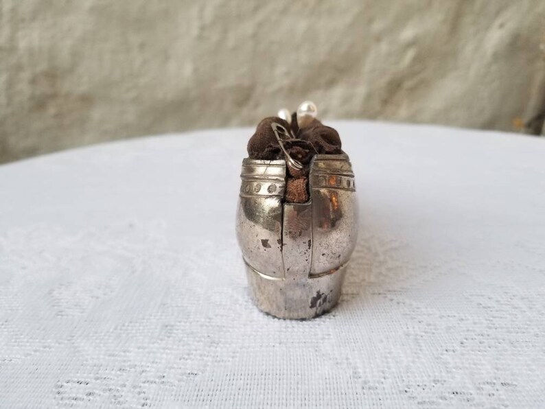 Pode incluir: Uma caixa de metal prateada em forma de sapato com um forro de tecido marrom. A caixa tem uma tampa com dobradi&ccedil;as e um design decorativo.