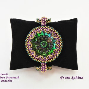 Könnte beinhalten: Ein grünes, goldenes und violettes Perlenarmband mit einem runden, schillernden grünen Mittelstück. Das Armband liegt auf einem schwarzen Samtkissen. Der Text "Nchantme® Jayashree Paramesh Illume Bracelet" und "Green Sphinx" ist unterhalb des Armbands sichtbar.